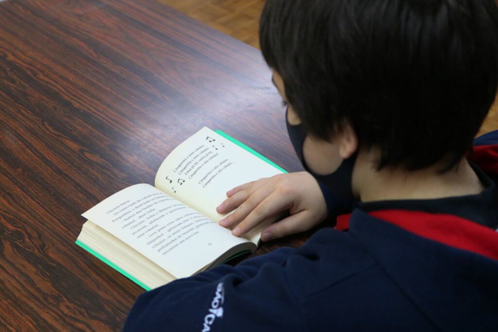 Menino uniformizado lendo livro posicionado em cima de uma mesa.